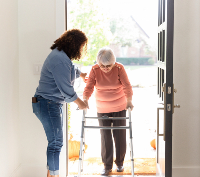 woman helping elderly woman with a crutch into a house